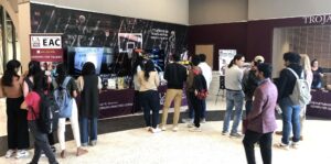 A crowd of people surrounds an exhibit table, speaking with representatives while viewing information on the exhibit screens and tablets.