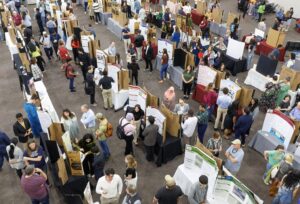 Large groups of people talk to presenters in front of presenter's presentation boards.